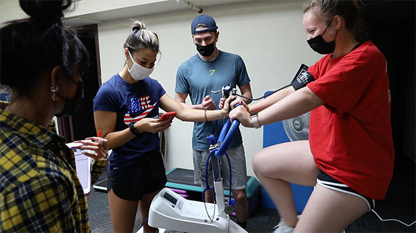 photo of a three students surrounding one riding an stationary bike