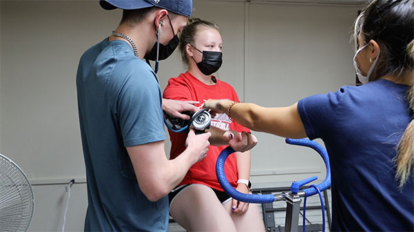 photo of a two students taking blood pressure reading from a young woman on a stationary bike