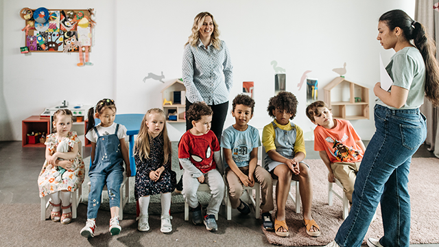 image with two teachers standing in front of a group of young students sitting in chairs